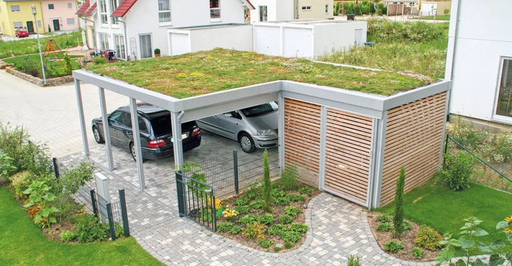Ein Carport mit begrüntem Dach schützt zwei geparkte Autos vor einem modernen Wohnhaus, umgeben von einem gepflegten Garten und gepflasterten Wegen in einer Vorstadtsiedlung.