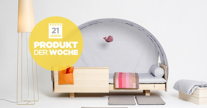 Galerie-Bild 1: Eine Holzkinderbett mit grauem Baldachin und rosa Kissen befindet sich neben einer Stehlampe auf einem hellen Hintergrund. Text auf gelbem Kreis: 21 PRODUKT DER WOCHE.