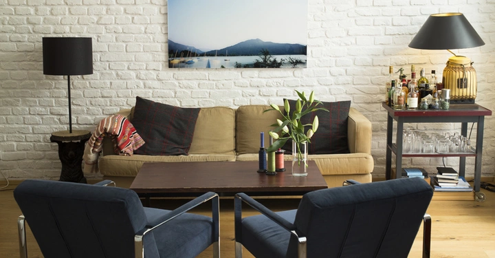 Galerie-Bild 3: Ein beiges Sofa steht vor einer weißen Ziegelwand mit einem Landschaftsbild. Zwei blaue Stühle, ein Couchtisch, eine Tischlampe, ein Barwagen und Dekorationen komplettieren den gemütlichen Wohnraum.