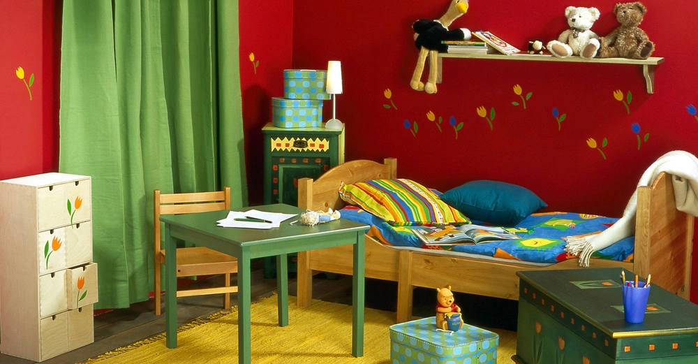 Holzbett mit bunter Bettwäsche steht in einem Kinderzimmer mit roten Wänden und grünen Vorhängen neben einem grünen Tisch und Stuhl zwischen bunten Spielzeug und Dekorationen über einen gelben Teppich.