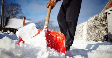 Eine Person schaufelt mit einer roten Schneeschaufel Schnee draußen vor einem Haus im Winter auf einem verschneiten Weg bei klarem Himmel und kahlen Bäumen.