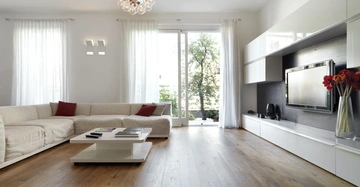 Ein großes weißes Sofa mit roten Kissen steht in einem modernen Wohnzimmer vor großen Fenstern mit weißen Vorhängen. Ein niedriger Couchtisch steht in der Mitte auf einem hellen Holzboden vor einem großen Fernseher und einem Schrank.