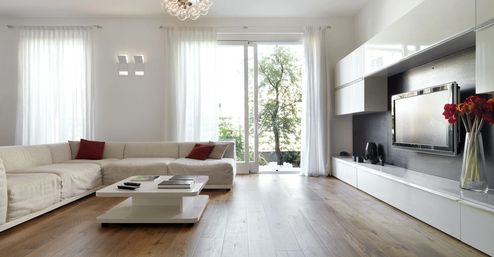 Ein großes weißes Sofa mit roten Kissen steht in einem modernen Wohnzimmer vor großen Fenstern mit weißen Vorhängen. Ein niedriger Couchtisch steht in der Mitte auf einem hellen Holzboden vor einem großen Fernseher und einem Schrank.