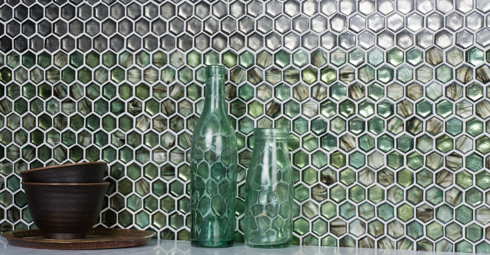 Braune Schalen und grüne Glasflaschen stehen auf einem Tisch vor einer Wand mit sechseckigen Mosaikfliesen und einem Farbverlauf von Grün zu Silber.