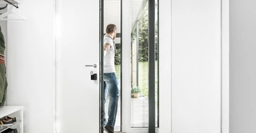Person öffnet eine weiße Tür und schaut nach draußen auf eine Terrasse mit Pflanzen. Umgebung ist ein heller Flur mit Holzboden und kleinem Schuhregal.