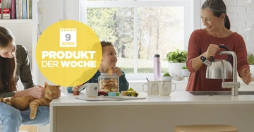 Eine Frau gießt Wasser aus einem Wasserkocher in einer modernen Küche ein während ein Kind am Tisch sitzend Plätzchen isst und eine andere Person daneben eine Katze streichelt. Text: 9 PRODUKT DER WOCHE