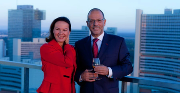 Galerie-Bild 23: Eine Frau und ein Mann lächeln und halten Weingläser auf einem Balkon, umgeben von hohen modernen Gebäuden bei Sonnenuntergang oder in der Abenddämmerung.