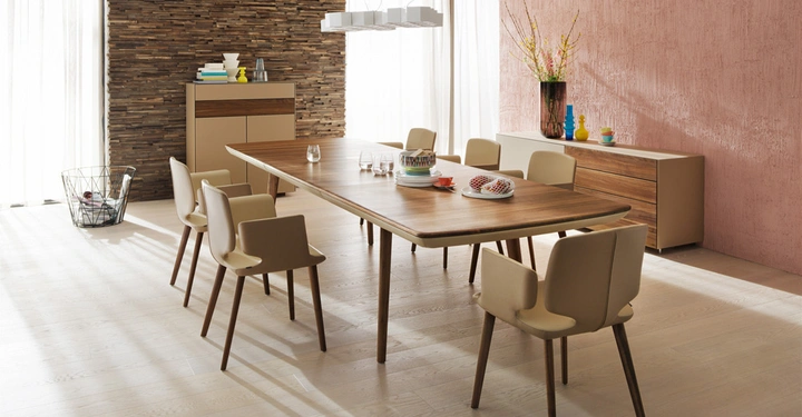 Galerie-Bild 10: Esszimmertisch aus Holz mit sechs beigefarbenen Stühlen in einem modernen Esszimmer mit Holzdielen und Steinwand sowie Sideboard mit Dekorationen im Hintergrund. Tageslicht durchflutet durch die Fenster.