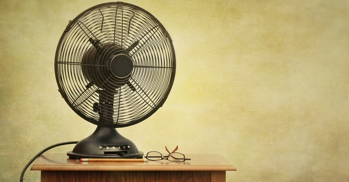 Tischventilator steht auf einem Holztisch vor einer beigen Wand links neben einer Brille und einem Stift.