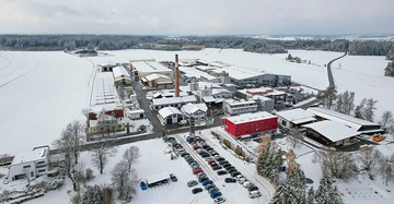 Gebäude stehen verschneit in einem Industriegebiet auf dem Land viele parkende Autos eine Straße windet sich durch die schneebedeckte Landschaft in deren Hintergrund sich ein Wald befindet