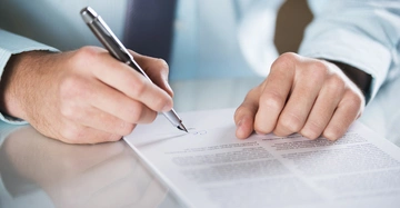 Eine Hand hält einen Stift und unterschreibt ein Dokument auf einem Tisch in einem Büro.