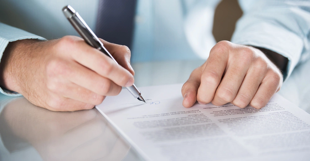Eine Hand hält einen Stift und unterschreibt ein Dokument auf einem Tisch in einem Büro.