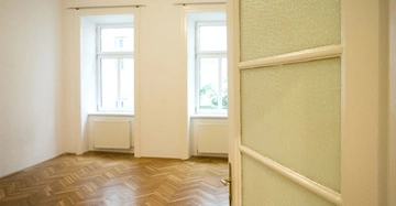 Tür öffnet sich zu leerem Zimmer mit Parkettboden und zwei großen Fenstern; weiße Wände und Heizkörper unter den Fenstern; helle und minimalistische Umgebung.