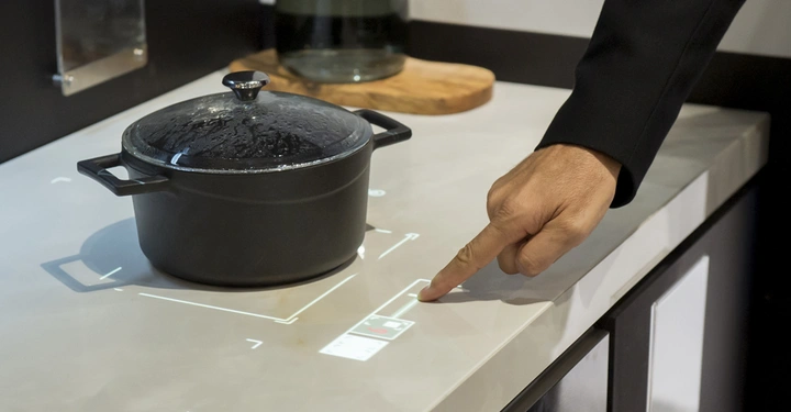 Ein schwarzer Topf steht auf einer Arbeitsplatte, während eine Hand auf ein projiziertes Bedienfeld zeigt. Im Hintergrund befindet sich ein Schneidebrett mit einem Glas mit Wasser.