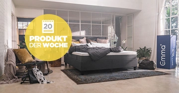 Ein ungemachtes Bett mit grauem Rahmen steht in einem hellen gemütlichen Schlafzimmer. Der Raum hat große Fenster Pflanzen und verschiedene Möbel. Text im Bild: "20 PRODUKT DER WOCHE" "Emma".