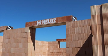 Baustelle mit unvollständigen Ziegelwänden unter blauem Himmel ein Stahlbalken mit der Aufschrift HELUZ spannt sich über eine Öffnung in einer Wand