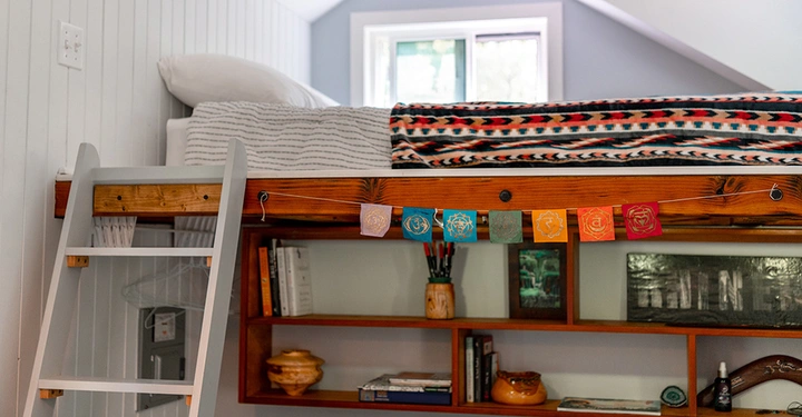 Leiter führt zu einem Hochbett geschmückt mit bunten Wimpeln vor einem Fenster unten Regal mit Büchern und Dekorationen in einem hellen Zimmer