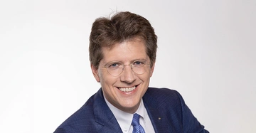 Michael Wardian mit randloser Brille und Lächeln im Gesicht. Portrait in dunkelblauem Anzug, weißem Hemd und lila Krawatte.