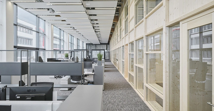 Innenaufnahme eines Großraumbüros, alles weiß und beige, hellgraue Teppichböden, Glasfronten zu den anderen Büros, große Fensterflächen links. Schreibtische und Sessel