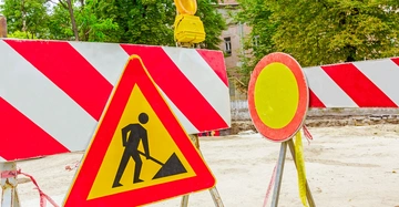 Baustellenschild zeigt arbeitende Person vor rot-weißen Absperrbaken auf Baustelle umgeben von Bäumen und Gebäuden