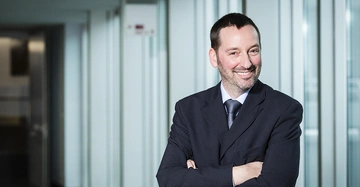 Mann lächelt und verschränkt die Arme im Anzug in einem modernen Bürogebäude mit Glaswänden und offener Tür im Hintergrund