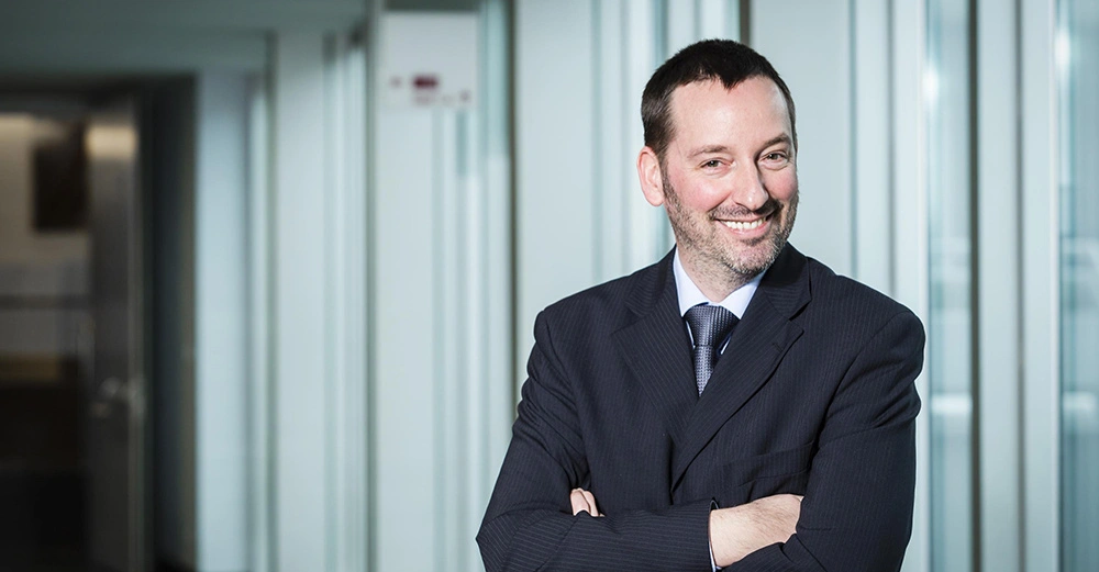 Mann lächelt und verschränkt die Arme im Anzug in einem modernen Bürogebäude mit Glaswänden und offener Tür im Hintergrund