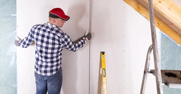 Eine Person befestigt eine Trockenbauplatte an einer Wand in einem Raum unter dem Dach mit Holzbalken und einer Aluminiumleiter