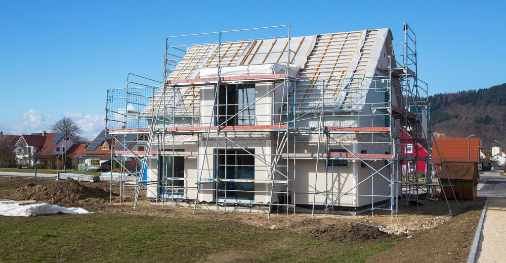 Ein zweistöckiges Haus wird gebaut das Gerüst umgibt das Gebäude in einem Vorort mit weiteren Häusern im Hintergrund unter klarem Himmel