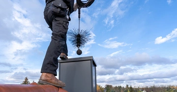 Rauchfangkehrer von Hüfte abwärts fotografiert steht auf Hausdach und reinigt den Kamin mit seinem Rauchfangwerkzeug. Blauer Himmel mit einigen Wolken im Hintergrund.