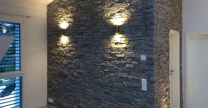 Steinwand mit eingeschalteten Wandleuchten in einem modernen, minimalistisch eingerichteten Raum mit weißen Wänden, einer Tür und einem Fenster mit Jalousien.
