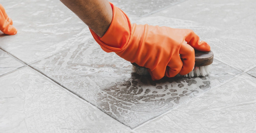 Hand in orangefarbenem Putzhandschu schrubbt Steinfliesen mit einer Bürste und Seifenwasser.