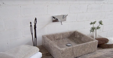 Rechteckiges Steinwaschbecken sitzt auf hölzernem Waschtisch vor weißer Ziegelwand Edelstahlarmatur darüber Handtücher links Dekorationsgegenstände und Pflanzentopf rechts großflächiger Spiegel darüber vermittelt minimalistischen Dekor