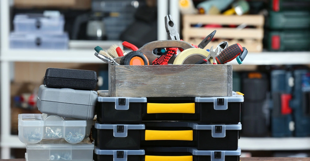 Auf einem Holztisch stehen mehrere schwarze Werkzeugkästen mit gelben Griffen gestapelt im Vordergrund und eine Holzkiste mit verschiedenen Werkzeugen darin im Zentrum in einem Regal mit weiteren Werkzeugen und Utensilien im Hintergrund.