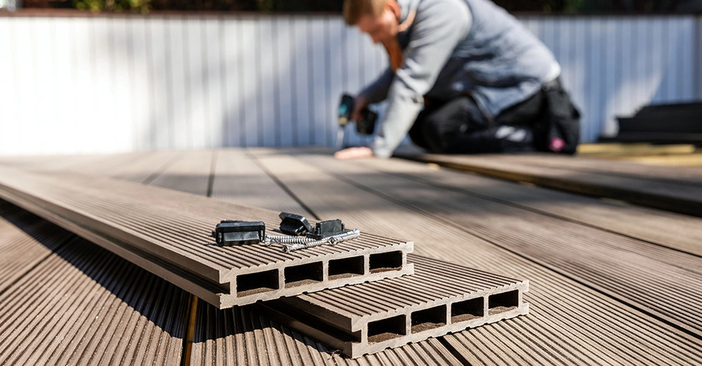 Montage von WPC Terrassendielen: Im Vordergrund liegen die Hohlkammerdielen und Befestigungsclips, während im Hintergrund ein Handwerker den Terrassenboden fachgerecht verlegt.