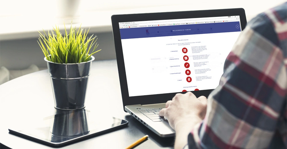 Person arbeitet an einem Laptop in einem geschäftigen Büroumfeld mit einer grünen Pflanze und einem Tablet auf dem Tisch.