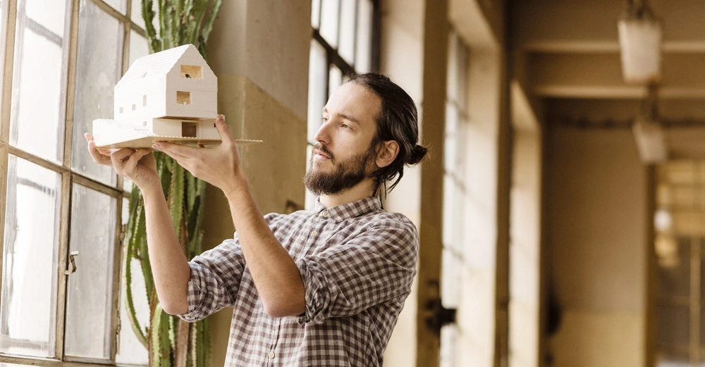 Ein Mann hält ein Modellhaus in den Händen und betrachtet es intensiv. Er steht in einem gut beleuchteten Raum mit großen Fenstern und einem Kaktus im Hintergrund.