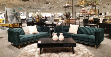 Grüne Sofas mit Kissen stehen vor einem schwarzen Couchtisch mit zwei Vasen in einem gut beleuchteten Möbelgeschäft, umgeben von Pflanzen Kunstwerken und weiteren Möbelstücken. Preisschilder sichtbar.