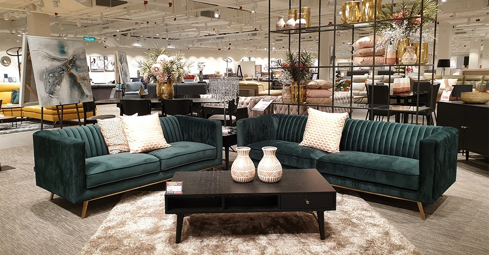 Grüne Sofas mit Kissen stehen vor einem schwarzen Couchtisch mit zwei Vasen in einem gut beleuchteten Möbelgeschäft, umgeben von Pflanzen Kunstwerken und weiteren Möbelstücken. Preisschilder sichtbar.