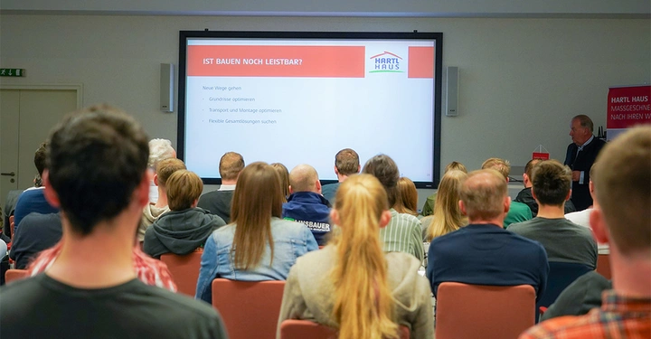 Personen sitzen in einem Seminarraum und hören einer Präsentation auf einer Leinwand zu. Die Leinwand zeigt den Text IST BAUEN NOCH LEISTBAR? HARTL HAUS. Neue Wege gehen Grundrisse optimieren Transport und Montage optimieren Flexible Gesamtlösungen suchen.