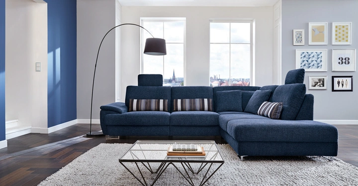 Ein blaues Sofa steht auf einem grauen Teppich in einem modern eingerichteten Wohnzimmer mit blauen und grauen Wänden; im Hintergrund ein großes Fenster und eine Bilderwand