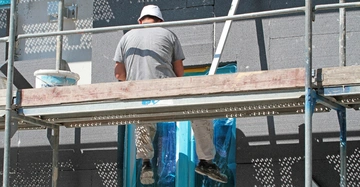 Maler sitzt auf Gerüst und klebt Dämmplatten an Hausfassade fest. Teil der Fassade ist schon fertig gedämmt