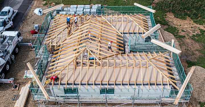 Galerie-Bild 3: Arbeiter bauen Holzdachkonstruktion auf einem Gebäude zusammen in Baugerüstumgebung, umgeben von Baufahrzeugen und Baumaterialien auf einer Kies- und Grasfläche.