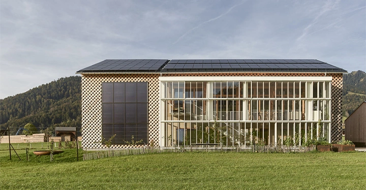 Zweistöckiges Gebäude mit Solarzellen auf dem Dach große verglaste Front mit Vertikallamellen in ländlicher Umgebung mit Wiesen und bewaldeten Hügeln im Hintergrund