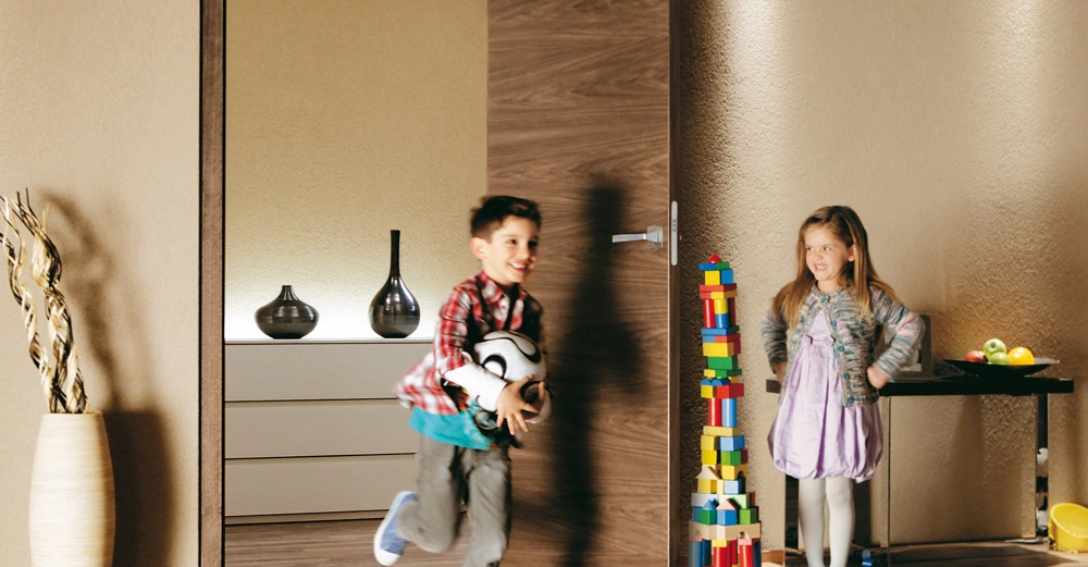 Junge läuft durch offene Tür mit Fußball im Arm, Mädchen steht neben hohem Turm aus Bauklötzen. Im Hintergrund moderne Möbel und Dekorationen in einem gut beleuchteten Raum.