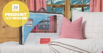 Verpackte Bettdecke befindet sich auf einem weißen Bett neben einem karierten Kissen in einem rustikalen Raum mit Holzverkleidung und Bergblick Text: 39 PRODUKT DER WOCHE sehr gut WELLNESS ZIRBE HEFEL