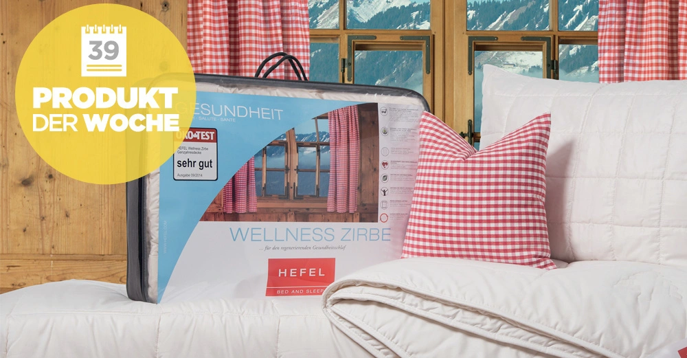 Verpackte Bettdecke befindet sich auf einem weißen Bett neben einem karierten Kissen in einem rustikalen Raum mit Holzverkleidung und Bergblick Text: 39 PRODUKT DER WOCHE sehr gut WELLNESS ZIRBE HEFEL