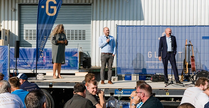 Drei Personen sprechen auf einer Bühne bei einer Veranstaltung im Freien vor einem Sitzpublikum Drei Banner mit der Aufschrift glorit.at sind sichtbar. Text: WIR BAUEN HIER FÜR SIE EXKLUSIVE HÄUSER Das 1. Haus am Platz
