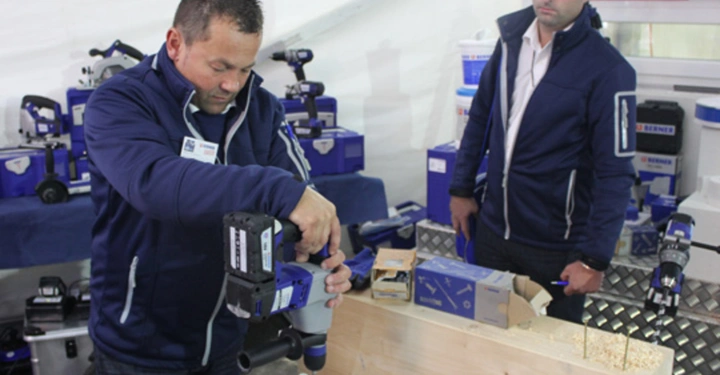 Galerie-Bild 12: Ein Mann bohrt ein großes Loch in ein Holzstück ein während ein weiterer Mann zusieht In einem Zelt sind verschiedene Elektrowerkzeuge und Kartons aufgestellt