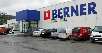 Autos parken vor einem weißen Gebäude auf einem nassen Parkplatz BERNER steht in großen blauen Buchstaben auf der Gebäudewand links ist ein gläserner Eingang rechts stehen mehrere Autos