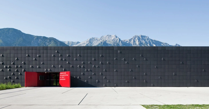 Galerie-Bild 7: Gebäude mit schwarzer Fassade und vielen dreidimensionalen geometrischen Mustern führt zu einem roten Eingang mit folgendem Text Schindler City Installation/Schindler Besucher Zentrum Kunsthalle Ziegelhütte mit Berglandschaft im Hintergrund und einem Betonvorplatz.